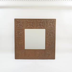 May include: Square, bronze-colored mirror with a decorative, embossed pattern on the frame. The frame has a repeating floral design. The mirror is centered within the frame and has a reflective surface.