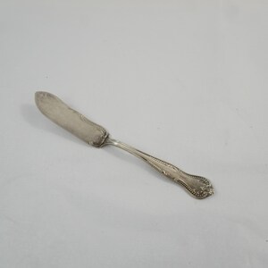May include: A silver-toned butter knife featuring a rounded, flat blade and a decorative handle with embossed bead and scroll detailing along the edges. The metal surface shows a polished finish with subtle signs of wear and tarnishing.