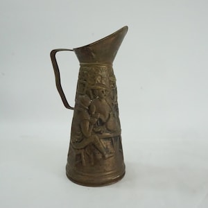 Könnte beinhalten: Eine Kanne aus Messing mit einem verzierten Design, das eine Szene von Menschen zeigt, die um einen Tisch sitzen. Die Kanne hat einen Henkel und einen Ausguss.