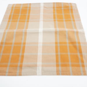 Op de afbeelding: Een gevouwen rechthoekige stoffen servet met een oranje en crème ruitpatroon. Het ruitpatroon bestaat uit oranje en crème vierkanten met dunne grijze lijnen. De servet ligt op een witte achtergrond.