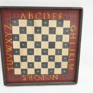 Puede incluir: Tablero de ajedrez cuadrado de madera con borde rojo oscuro y marco negro. El tablero tiene cuadrados negros y beige alternados. Letras y corazones dorados decoran el borde, con el alfabeto alrededor de los bordes.