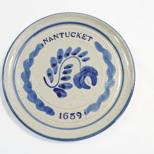 Puede incluir: Plato redondo de cerámica con base crema y detalles azules. El plato presenta la palabra "NANTUCKET" en azul, junto con un diseño floral y el número "1659". El borde tiene un borde azul ondulado.