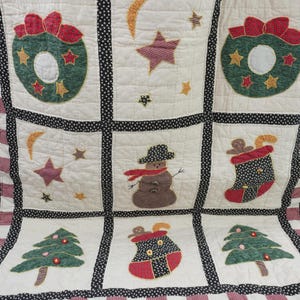 May include: A quilted throw featuring a grid of nine squares with Christmas motifs including green wreaths with red bows, a brown snowman with a scarf, red and black stockings, and green pine trees, all bordered by black polka dot and red striped fabric.
