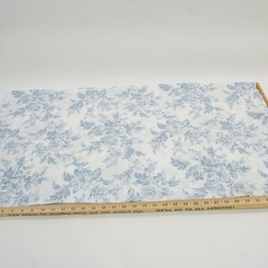 May include: White fabric with a blue floral pattern. A wooden ruler is placed across the fabric. The fabric is likely used for sewing or crafting projects. The floral design features detailed leaves and flowers.