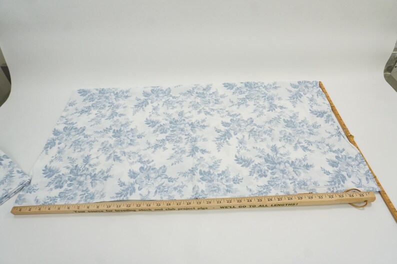 May include: White fabric with a blue floral pattern. A wooden ruler is placed across the fabric. The fabric is likely used for sewing or crafting projects. The floral design features detailed leaves and flowers.
