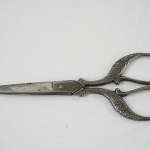 Könnte beinhalten: Eine Metallschere mit dunklem, verwittertem Finish. Die Griffe weisen ein kompliziertes, geprägtes Blumen- und Geometriemuster auf, während die Klingen gerade und spitz sind und Gebrauchsspuren sowie leichte Verfärbungen auf der Oberfläche aufweisen.