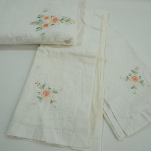 May include: White embroidered linens featuring a floral design with pink and green accents. The linens are folded and arranged, showcasing the delicate embroidery. The overall theme is vintage bedding.