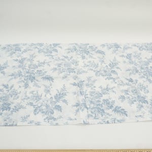 May include: A rectangular piece of fabric with a white background and a blue floral pattern. The pattern features detailed illustrations of flowers and foliage. A wooden ruler is at the bottom of the image.