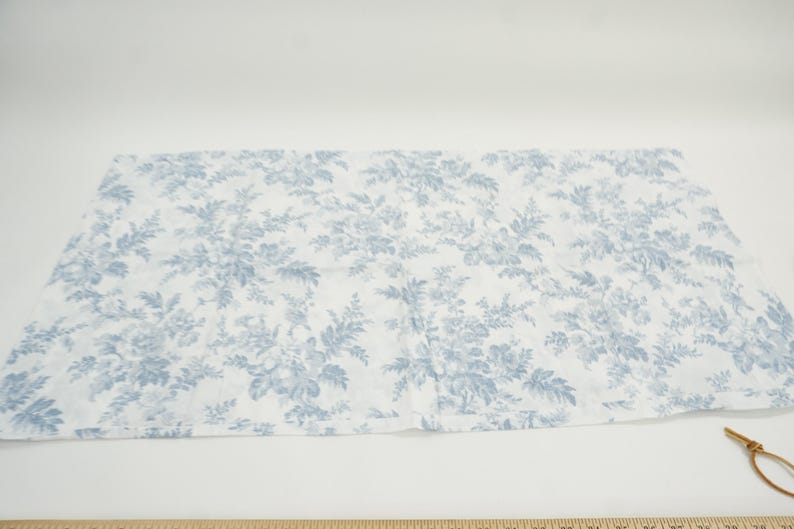 May include: A rectangular piece of fabric with a white background and a blue floral pattern. The pattern features detailed illustrations of flowers and foliage. A wooden ruler is at the bottom of the image.