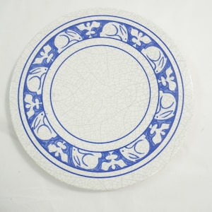May include: A round, off-white ceramic plate with a blue border featuring a repeating pattern of rabbits and floral elements. The plate has a cracked, aged appearance, and the center is plain.