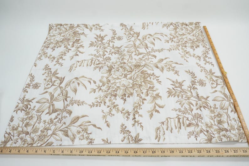 May include: White fabric with a brown floral pattern. A wooden ruler is placed along the edge of the fabric. The fabric features a repeating design of flowers and foliage, creating a classic, elegant look. The ruler measures up to 28 inches.