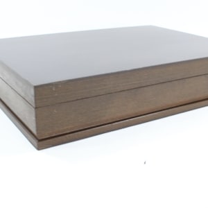Puede incluir: Una caja de madera marrón oscuro con una tapa con bisagras. La caja está cerrada y se encuentra sobre una superficie blanca.