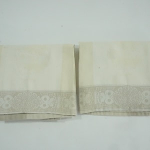 May include: Two white fabric napkins with a decorative border of a repeating pattern in a light gray color.