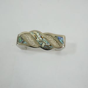 Könnte beinhalten: Silberfarbenes Armband mit geflochtenem Design. Die Mitte zeigt schillernde Abalone-Muschel und funkelnde Akzente. Das Armband ist etwa 2,5 cm breit.