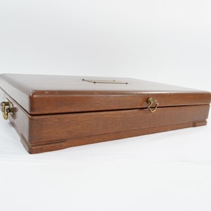 May include: A rectangular wooden box with a smooth, dark brown finish. The box features a hinged lid with a small, centered metal plate on top, a brass-colored latch on the front, and a decorative brass-colored handle on the side.