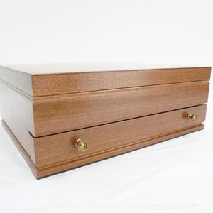 Puede incluir: Una caja de joyería de madera con un acabado marrón cálido. La caja tiene forma rectangular con un cajón y una tapa. El cajón tiene un pomo redondo de color dorado. La caja está sobre un fondo blanco.