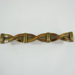 Könnte beinhalten: Dekorative Spangenbrosche mit geflochtenem Design. Die Brosche hat einen goldfarbenen Metallrahmen mit einem zentralen Muster aus verschlungenen braunen und beigen Abschnitten. Die Enden der Brosche sind ebenfalls goldfarben.