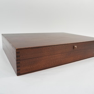 May include: A rectangular wooden box with a dark brown finish and visible finger joint construction on the side panels. The lid features a small, centered square knob for opening, showcasing a smooth, flat surface with natural wood grain.