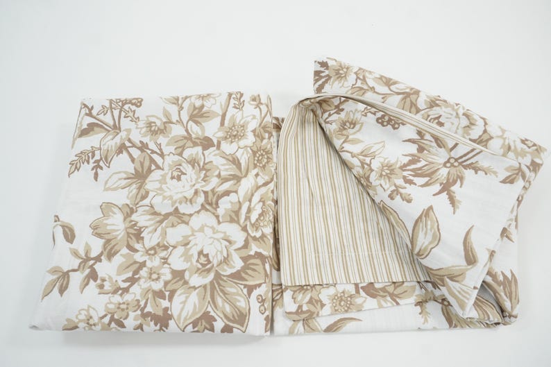 May include: A folded floral fabric with a cream background and large beige flowers and leaves. The reverse side features a beige and white striped pattern. The fabric is likely for bedding or home decor.