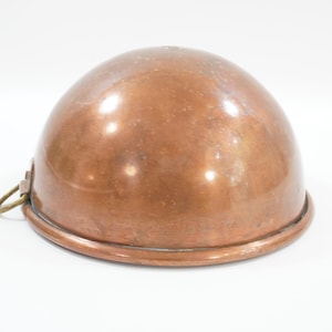 May include: A copper-colored, dome-shaped bowl with a rounded base and a small metal loop attached to the side. The bowl has a shiny, reflective surface. The item appears to be a kitchenware item.