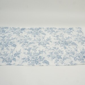 May include: A white fabric with a blue floral pattern. The pattern features detailed flowers and foliage, creating a classic, elegant design. The fabric is spread out on a white surface.