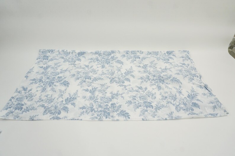 May include: A white fabric with a blue floral pattern. The pattern features detailed flowers and foliage, creating a classic, elegant design. The fabric is spread out on a white surface.
