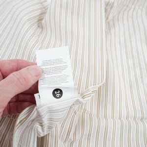 May include: Close-up of a cream-colored fabric with vertical brown stripes. A white tag with black text and the number 26 is attached to the fabric. The fabric is being held by a person.