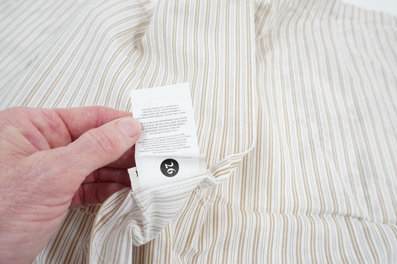 May include: Close-up of a cream-colored fabric with vertical brown stripes. A white tag with black text and the number 26 is attached to the fabric. The fabric is being held by a person.