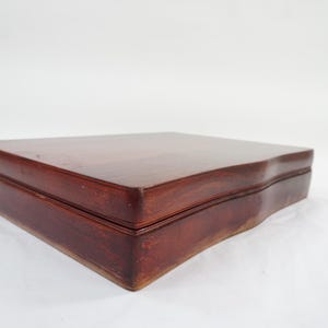 Puede incluir: Una caja rectangular de madera con un acabado liso en color marrón rojizo oscuro. La caja presenta una tapa plana y un borde frontal ligeramente curvado, mostrando un patrón de veta de madera natural en toda la superficie del contenedor.