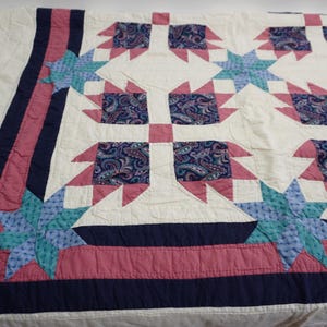 May include: A handmade quilt with a repeating pattern of white, pink, blue, and navy blue shapes. The quilt features a central design with paisley fabric accents, surrounded by star and geometric patterns. The border is navy blue and pink.