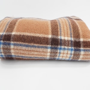 May include: A folded fleece blanket featuring a plaid pattern in shades of tan, brown, cream, and thin blue stripes. The fabric has a soft, textured appearance with a consistent grid design across the entire surface of the folded textile.