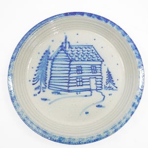 May include: Round ceramic plate with a blue and white cabin design. The plate features a blue cabin, trees, and a path, all set against a white background. The rim has a blue border.