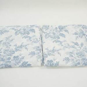 May include: Two folded white fabric pieces with a blue floral pattern. The pattern features detailed blue flowers and foliage against a white background. The fabric appears to be soft and lightweight, suitable for bedding or curtains.