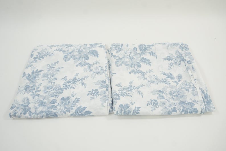 May include: Two folded white fabric pieces with a blue floral pattern. The pattern features detailed blue flowers and foliage against a white background. The fabric appears to be soft and lightweight, suitable for bedding or curtains.