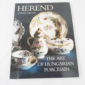 Puede incluir: Una portada de libro con porcelana Herend. La portada muestra una variedad de piezas de porcelana, incluyendo una tetera, un plato con tapa, una taza y un plato. Las piezas son blancas con diseños florales y de mariposas. El texto dice "The Art of Hungarian Porcelain."
