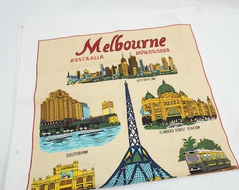 Puede incluir: Un paño de cocina de recuerdo color crema con Melbourne, Australia. El paño tiene un borde rojo e incluye ilustraciones del horizonte de la ciudad, la estación Flinders Street y otros puntos de referencia. El texto incluye "Melbourne" y "Downunder".