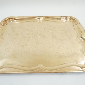 May include: A rectangular brass tray with rounded corners and a raised, scalloped edge. The surface of the tray features a subtle, etched floral pattern across the center, with a polished metallic finish that reflects light across its surface.