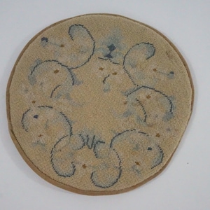 May include: A round, beige needlepoint coaster with a brown border. The coaster features a repeating pattern of stylized blue and brown floral or abstract shapes. The word "OUT" is visible in the design.
