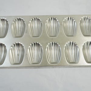 May include: A rectangular, silver madeleine pan with 12 shell-shaped moulds. Each mould has a fluted design. The pan is made of metal and is designed for baking small cakes.