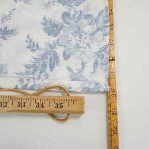 May include: White fabric with a blue floral pattern, measured with a wooden ruler. The ruler shows measurements in inches. A leather cord is attached to the fabric and ruler.