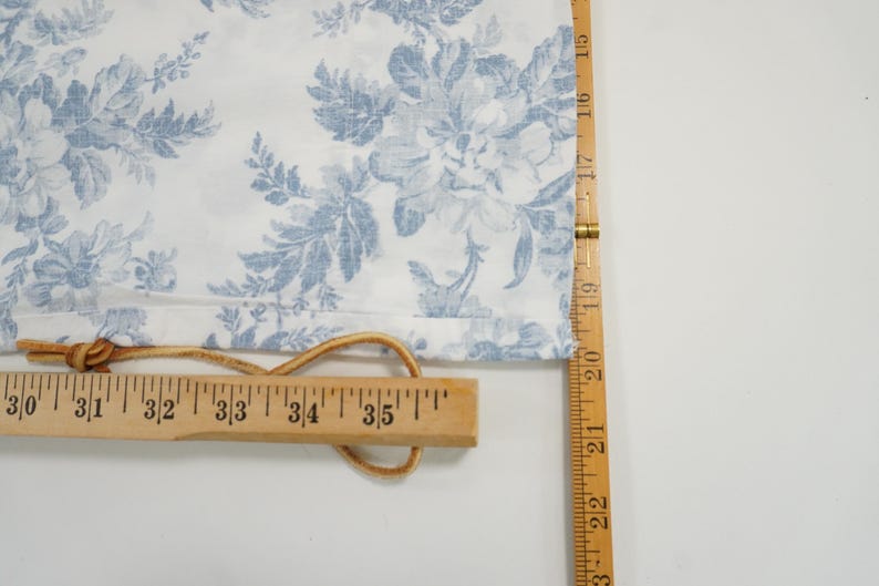 May include: White fabric with a blue floral pattern, measured with a wooden ruler. The ruler shows measurements in inches. A leather cord is attached to the fabric and ruler.