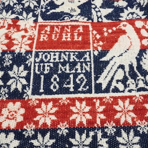 May include: Close-up of a woven textile with a repeating pattern in red, white, and navy blue. The design includes floral motifs, birds, and text that reads "ANNA RUHL" and "JOHNKA UF MAN 1842". The edges have cream-coloured fringe.