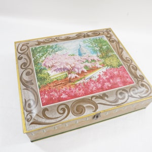May include: A rectangular, vintage metal box with a hinged lid. The lid features a framed image of a park scene with a building, trees, and pink flowers. The frame has a gold border and decorative scrollwork.