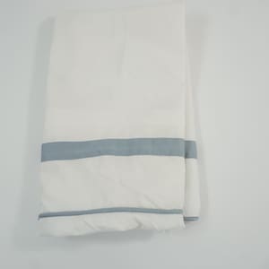 May include: A folded white textile item featuring two horizontal light blue stripes. The fabric has a smooth texture and a clean appearance, with one stripe positioned near the bottom edge and a second stripe located slightly higher up the surface.