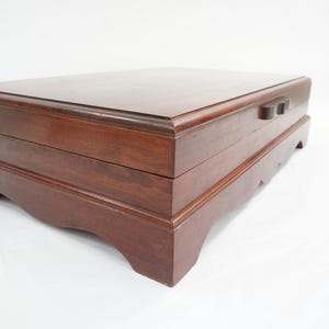 Puede incluir: Caja de joyería rectangular de madera marrón oscuro. La caja tiene una tapa con bisagras y un cierre de dos pomos. La base presenta patas curvas. La caja se encuentra sobre un fondo blanco.