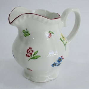 Puede incluir: Jarra de cerámica blanca con borde ondulado y asa. La jarra está decorada con diseños florales y de tréboles de colores. El borde tiene un borde rojo. Una pieza decorativa para la cocina o el comedor.