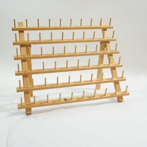 以下が含まれることがあります： 複数のピンが並んだ木製の糸巻きラック。明るい色の木材で作られたラックは、多数の糸巻きを収納できるように設計されており、縫製や手芸プロジェクトに最適です。