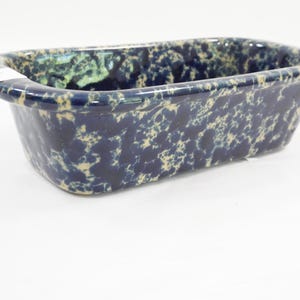 May include: A rectangular loaf pan with a glossy finish, featuring a dark navy blue and cream-colored speckled pattern throughout the exterior and interior surfaces. The bakeware has a wide, rounded rim and a deep, open basin design.