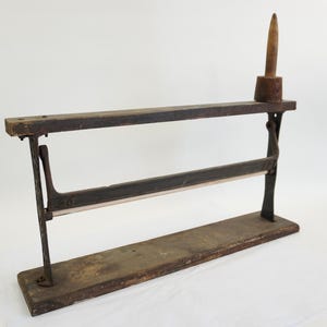 Puede incluir: Un cortador de papel antiguo de madera y metal. Tiene una base rectangular, dos barras horizontales y un soporte vertical. Un mango de madera está unido a la barra superior. Los componentes metálicos muestran signos de edad y desgaste.
