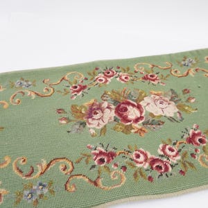 May include: A sage green needlepoint runner featuring a central floral arrangement of pink and cream roses surrounded by scrolling gold and brown filigree borders. The textile displays a repeating pattern of smaller rose clusters and foliage.